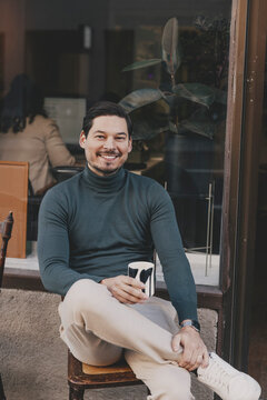 Portrait of smiling businessman holding coffee cup while sitting on chair outside office