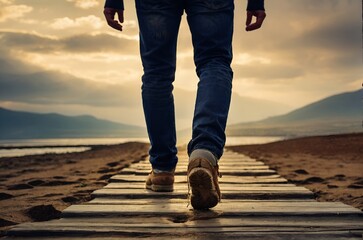 Person walking on a wooden beachside pathway under a cloudy mountain backdrop. Back view.