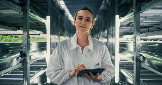 Portrait of a Female Agricultural Engineer Posing for Camera, Smiling. Specialist Standing in a Vertical Farm Facility Next to Racks with Fresh Natural Crops Illuminating with UV LED Light