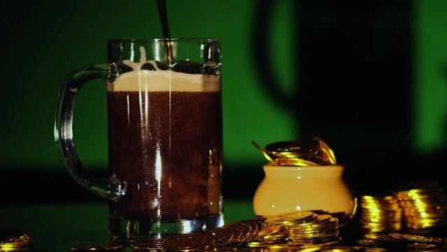 Fresh dark beer is pouring and foaming in glass on green background with pot of golden coins. St Patrick's Day concept. Video 4K. Copy space.
