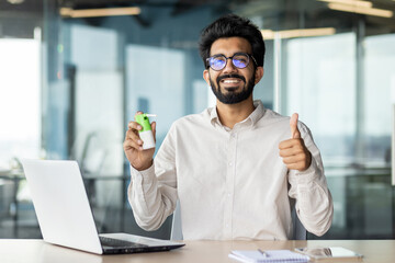 Portrait of a smiling Indian man sitting at a desk in the office, holding a throat spray in his...
