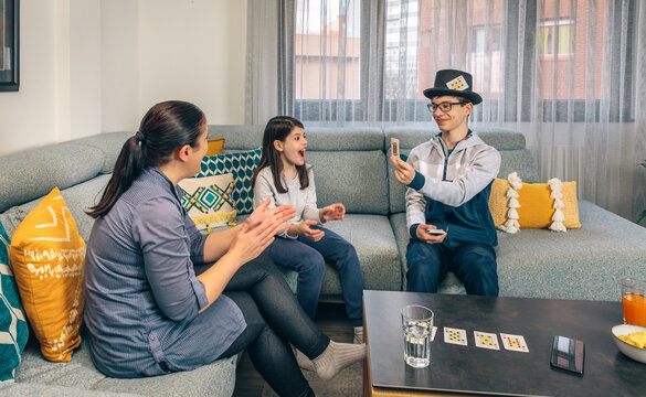 A family enjoys a card game together; a boy in a hat shows a card with excitement while a girl reacts with joy and a woman claps