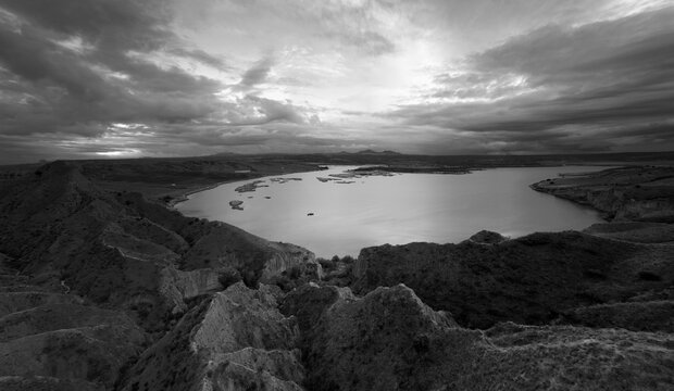 Expansive black and white landscape with dramatic clouds over a calm lake and rolling hills