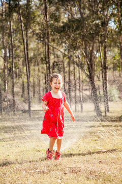 Happy little girl in pink dress laughing and running under sprinkler in backyard