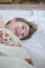 Obraz premium Child toddler boy is wrapped in a blanket like a roll, his head sticks out of the blanket. He is laughing, having fun on the bed. 