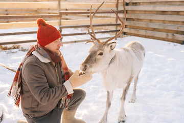 Portrait of handsome young man in warm clothes hugging and feeding cute young reindeer on snowy...