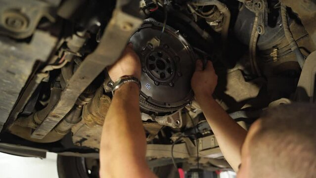 Auto mechanic replacing clutch in car. Hands remove old and install new clutch disc, cover, release bearing. Auto repair under lifted vehicle, workshop maintenance, transmission servicing.