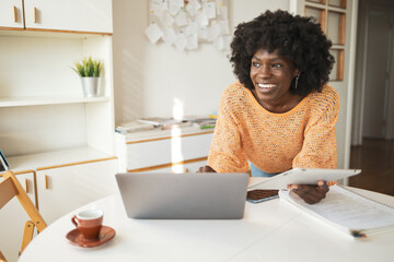 Happy thoughtful woman using laptop and digital tablet at table in home office