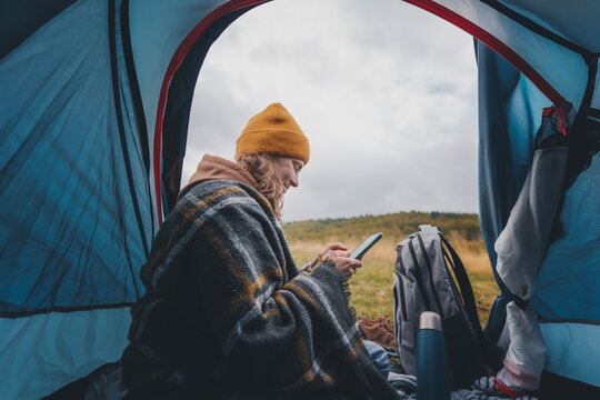 Young cheerful woman sitting in a tent and using smartphone while travelling. Hiking travel and trekking concept