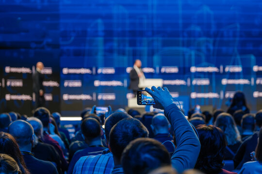 Business event attendees recording speaker on stage giving a professional presentation.
