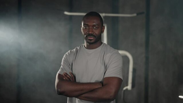 African american sportsman in gym. Man in white t-shirt smiling standing in close pose with hands crossed on chest ready for sport training. Healthy lifestyle, useful habits, strong body concept.