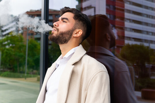 A contemplative businessman exhales smoke while standing thoughtfully near a reflective glass building, dressed in a stylish beige coat