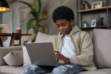 Young ethnic African American boy teenager uses laptop to play games on Internet sits on sofa with portable computer on lap. Digital addiction