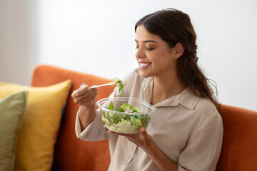 Relaxed young woman eating fresh green salad from bowl at home