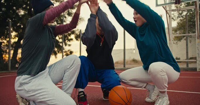 A trio of happy blonde girls on the same basketball team put their hands together and raise them up in a sign of unity near the basketball ball on the red basketball court early in the morning