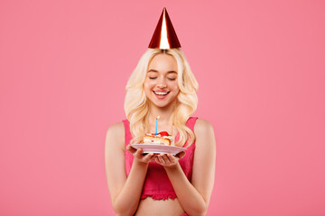Blissful young woman with birthday cake and party hat, making wish