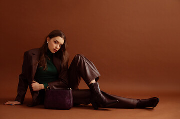 Fashionable confident woman wearing faux leather suit blazer, pants, pointed toe ankle boots, with trendy purple bag, posing on brown background. Full-length studio fashion portrait. Copy, empty space