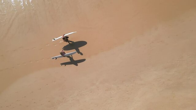 Aerial top view of surfers couple walking on sandy beach with surfboards by ocean waves. Drone flying over two people carrying boards along sea surf. View from above of young man, woman after session.