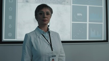 Portrait of female doctor wearing white coat posing for camera in front of projection screen with microscope images and graphs during medical conference