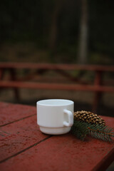a white cup stands on a red wooden table in the forest. a pine cone and fir branches are nearby
