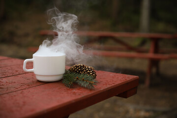 a white cup stands on a red wooden table in the forest. steam comes out of the cup. a pine cone and spruce branches are nearby