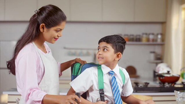 Caring Indian mother placing tiffin box and water bottle into kids backpack for going to school - concept of motherhood, parental caring and responsibility