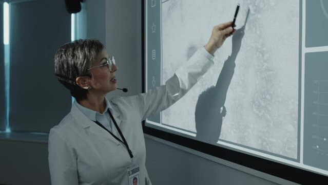 Female scientist wearing white coat and microphone headset pointing at microscope image and graph on projection screen, commenting on medical research and giving presentation on conference