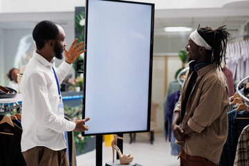 African american employee using smart display to assist customer with shoe purchase in shopping center. Clothing store showing promotion on whiteboard, helping customers selecting stiletto