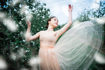 A beautiful girl in an airy long dress dances among a blooming apple orchard. Spring. Flying apple flower petals