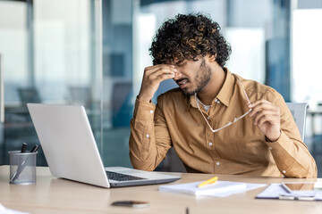 Indian businessman in casual shirt appearing stressed, sitting at his office desk with laptop,...