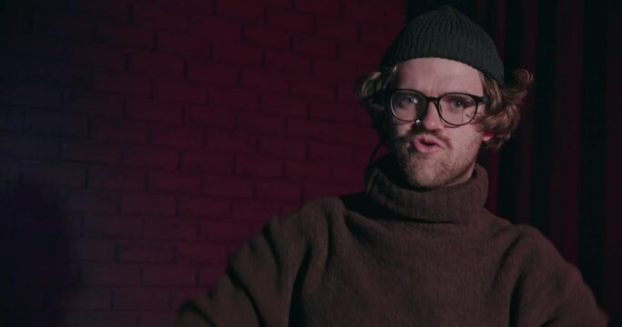 Positive funny guy in turtleneck training his muscles at studio on gradient light background. Humorous comic with glasses exercising with barbells. Stand up show about weak nerdy sports man