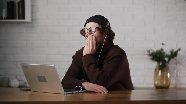 A young bored man listening lecture via laptop. Unmotivated employee feel sleepy during daily meet with team. Man listens a boring lecture at home using notebook. Uninteresting seminar online