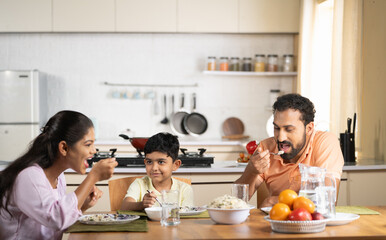 Happy parents with child eating lunch together on dining table at home - concept of weekend relaxation, family bonding and parenthood - obrazy, fototapety, plakaty