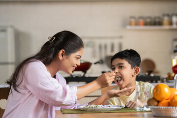 Happy Indian mother feeding to child at home on dining table - concept of parental caring, family bonding and motherhood.