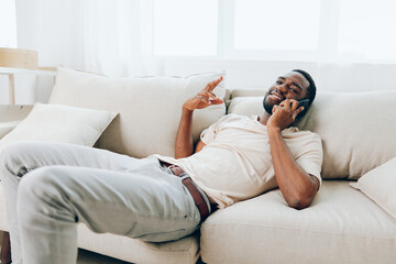 Happy African American man sitting on a black sofa, holding a smartphone and having a video call He...