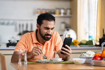 Indian middle aged man busy using mobile phone while eating lunch on dining table at home - concept of modern lifestyles, internet distraction and social media sharing - obrazy, fototapety, plakaty