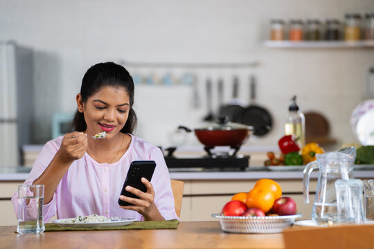 Indian young woman using mobile phone while eating food at home - concept of social media addiction, cyberspace and surfing internet