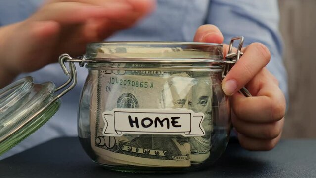 Unrecognizable woman add money to Saving Money In Glass Jar filled with Dollars banknotes. HOME transcription in front of jar. Managing personal finances extra income for future insecurity background