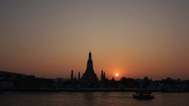 sunset provides a stunning backdrop to the iconic Wat Arun Temple along the Chao Phraya River in Bangkok, with silhouette and reflection.