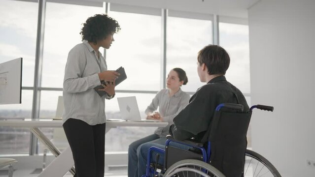 Paralyzed man works with colleagues. Guy in wheelchair has spine trauma discussing project in office on meeting with multiethnic team women colleagues. Teamwork brainstorming, special needs concept.