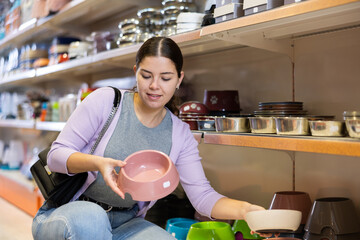 Interested young woman choosing functional and safe bowl for puppy in pet supplies store, looking and comparing different models
