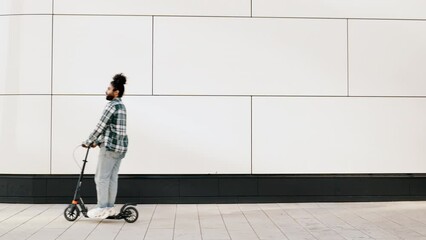Trendy smiling bearded man in casual clothes riding electric scooter in urban background. Handsome model posing in the street. Hipster guy with curly hairstyle. Cheerful and happy
