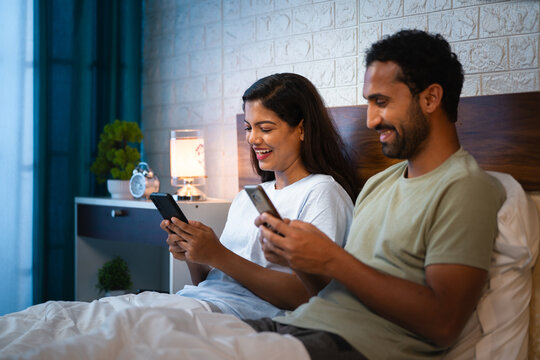 happy Indian couples busy using mobile phones on bedroom before sleeping at night - concept of social media or internet addiction and digital distraction