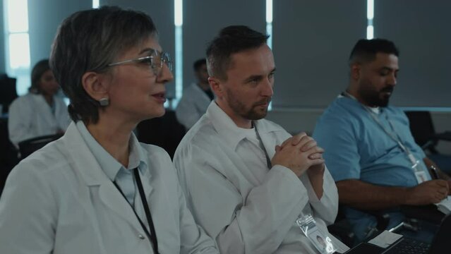 Female doctor speaking with male colleague while sitting together during medical conference and listening to presentation