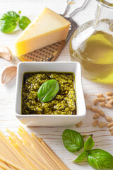 Homemade pesto sauce in small white square jar and ingredients for pasta on white wooden background. Traditional Italian cuisine, recipe, restaurant menu