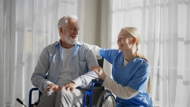 Nurse talking senior man in wheelchair. Female social welfare physical therapist looks after senior male, listening, providing medical care at home. Living with legs paralysis, special needs patient.
