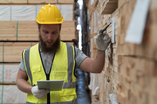 Male warehouse worker using digital tablet checks stock inventory in lumber storage warehouse. Male worker checks barcodes on pile of plank wooden in wooden warehouse