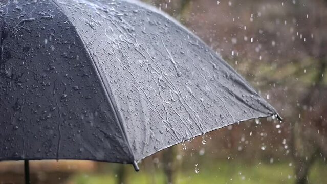 An outdoor black umbrella faces the rain, with water droplets scattering in slow motion, a gloomy day with sparse sunlight.