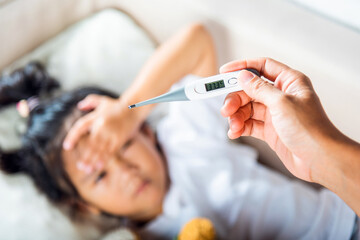 Obraz na płótnie Canvas Sick kid. Mother parent checking temperature of her sick daughter with digital thermometer in mouth, child laying in bed taking measuring her temperature for fever and illness, healthcare