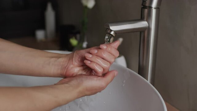 Close-up shot of bare hands of anonymous young woman lifting tap on bathroom faucet and washing hands under running water in white ceramic sink at home